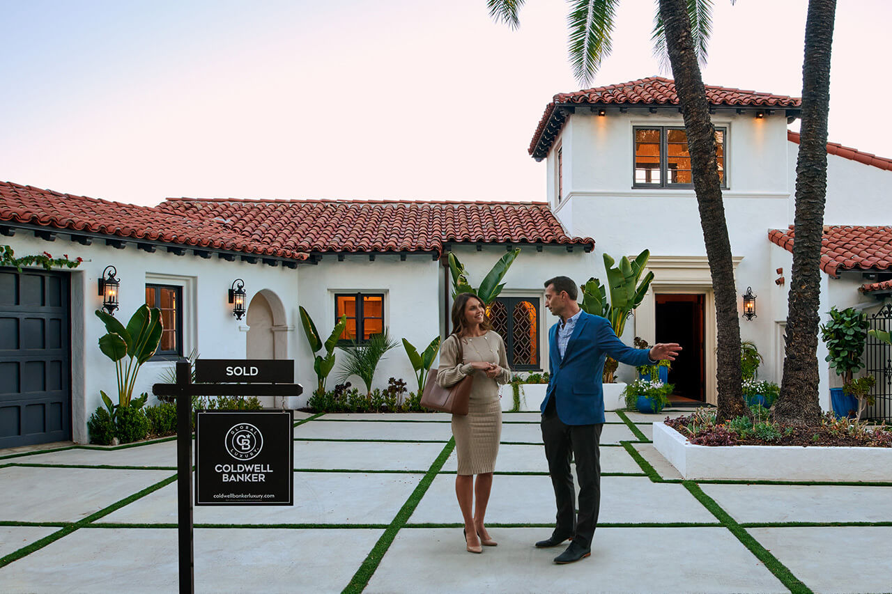 A smiling man and woman converse in front of a white Spanish-style house with a 'SOLD' sign by Coldwell Banker, indicating a successful real estate sale